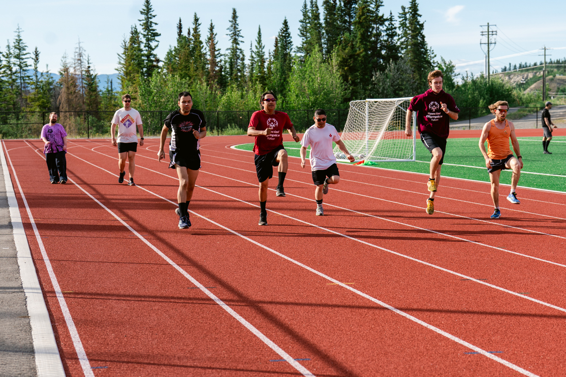 Seven athletes running along a track. It's a sunny day and the athletes are all wearing athleisure.