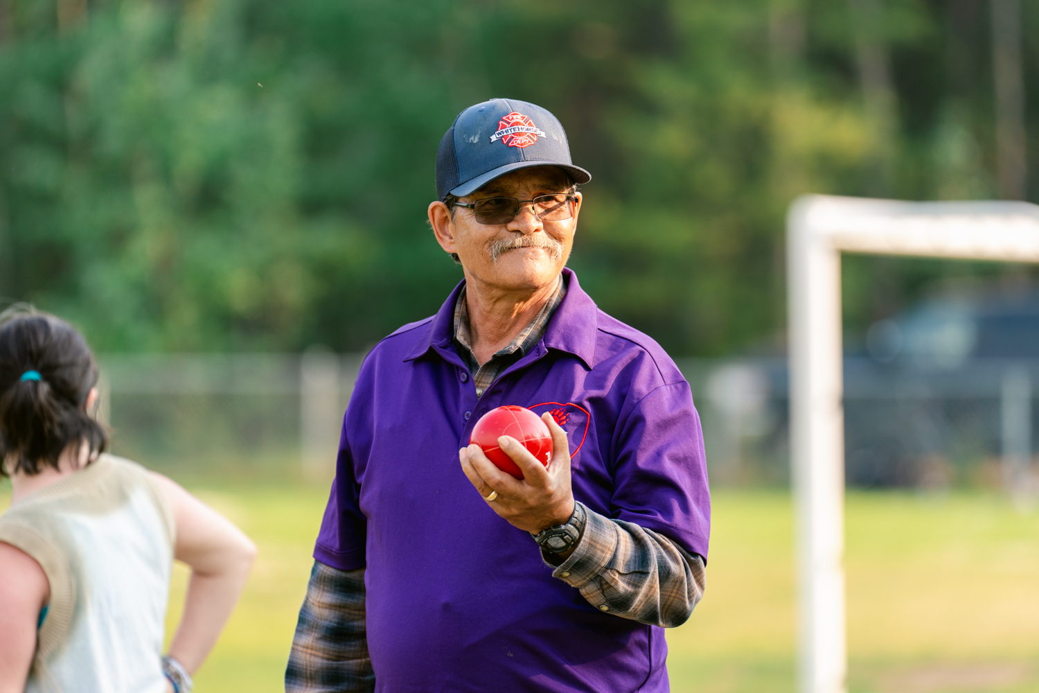 A close up image of a bocce player. He is wearing a purple collared shirt with a baseball cap and a pair of glasses. He is holding up a red bocce ball and staring into the distance smiling. A close up image of a bocce player. He is wearing a purple collared shirt with a baseball cap and a pair of glasses. He is holding up a red bocce ball and staring into the distance smiling.