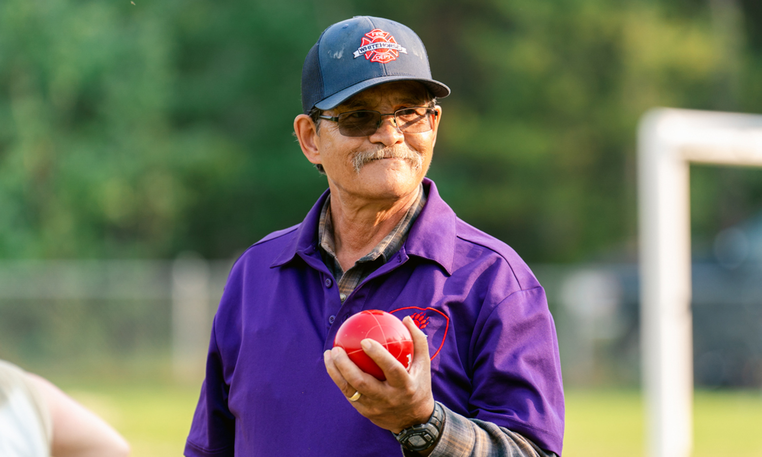 A close up image of a bocce player. He is wearing a purple collared shirt with a baseball cap and a pair of glasses. He is holding up a red bocce ball and staring into the distance smiling.