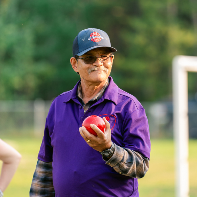 A close up image of a bocce player. He is wearing a purple collared shirt with a baseball cap and a pair of glasses. He is holding up a red bocce ball and staring into the distance smiling.