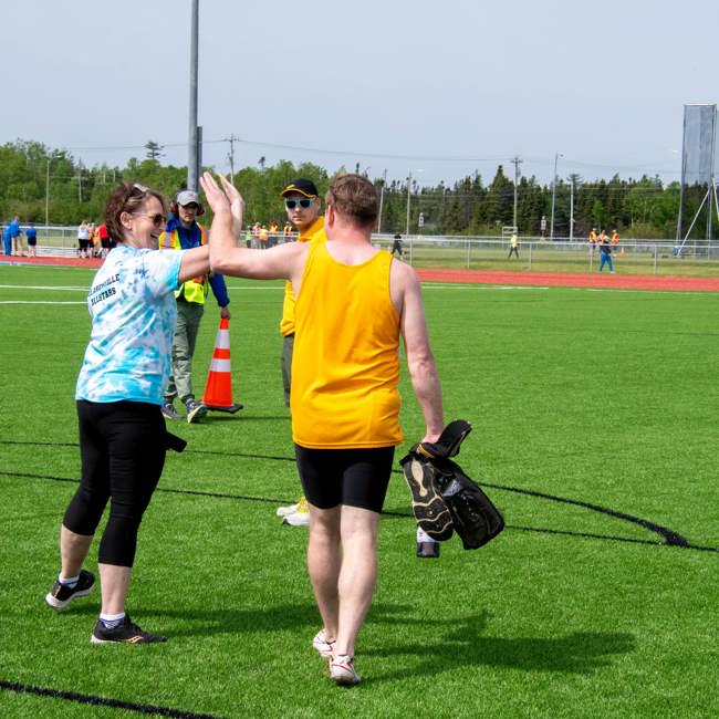 A coach is giving a high-five to an athlete. The photo was taken from behind the pair and you can only see a glimpse of the coach's face, smiling at the athlete. The athlete is holding his equipment in the hand not being used for the high five.