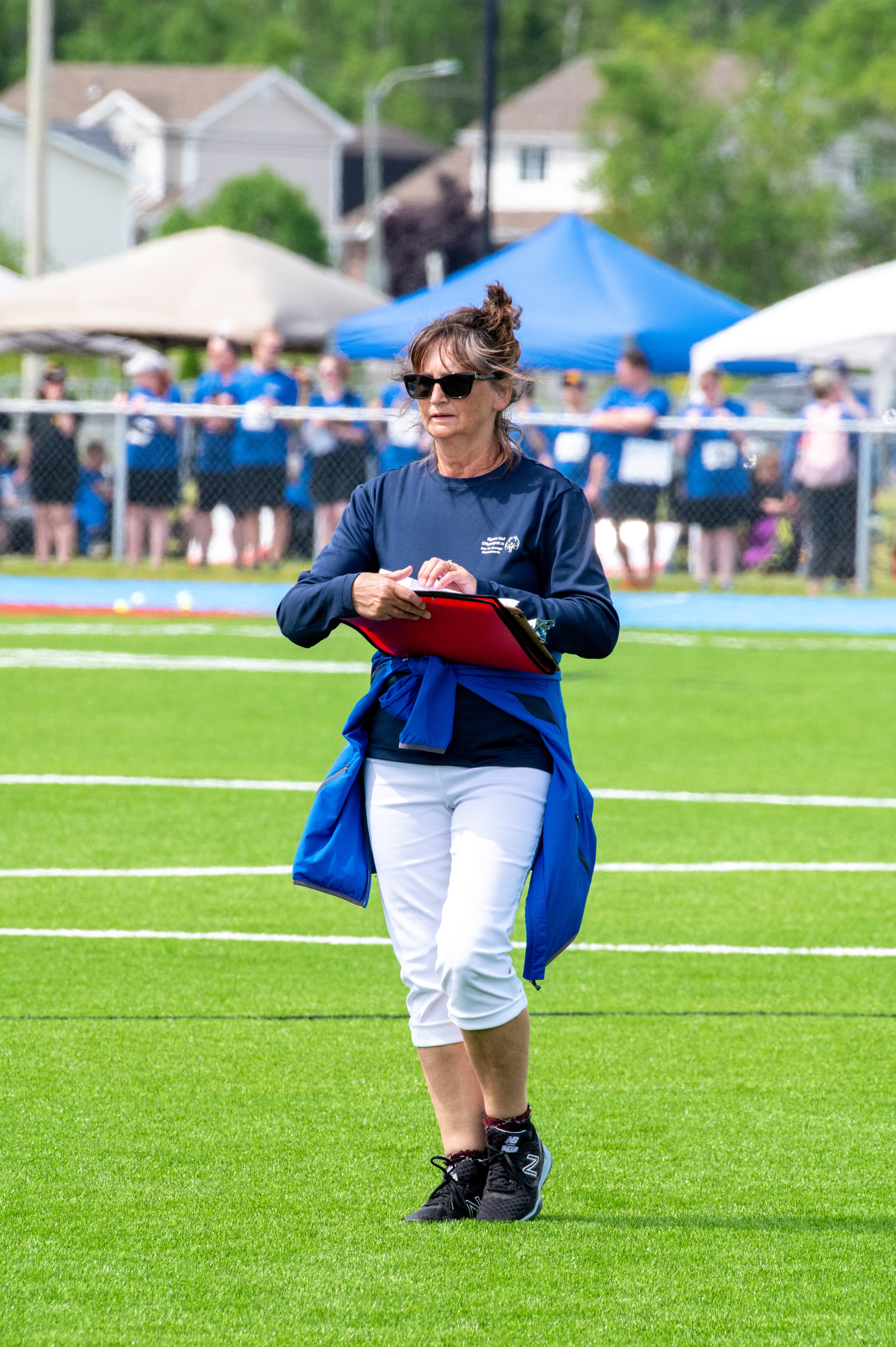 A volunteer with her red binder, is standing in the middle of the field. She is wearing sunglasses and a navy blue long sleeve shirt. She is wearing a light blue jacket tied around her waist.