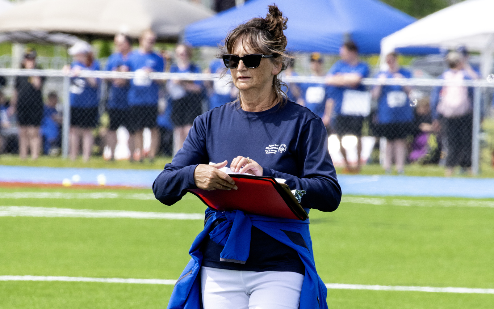 A volunteer with her red binder, is standing in the middle of the field. She is wearing sunglasses and a navy blue long sleeve shirt. She is wearing a light blue jacket tied around her waist.