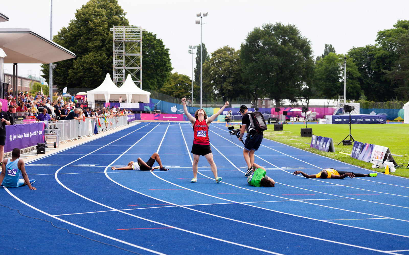 A triumphant Special Olympics BC athlete at the end of their race with their arms raised as fellow competitors lie exhausted on the track around him. 