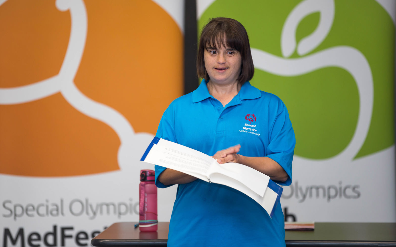 An athlete standing up and presenting a speech. She is wearing a blue Special Olympics Athlete Leadership collared shirt, and is holding a folder with her papers and speaker notes.