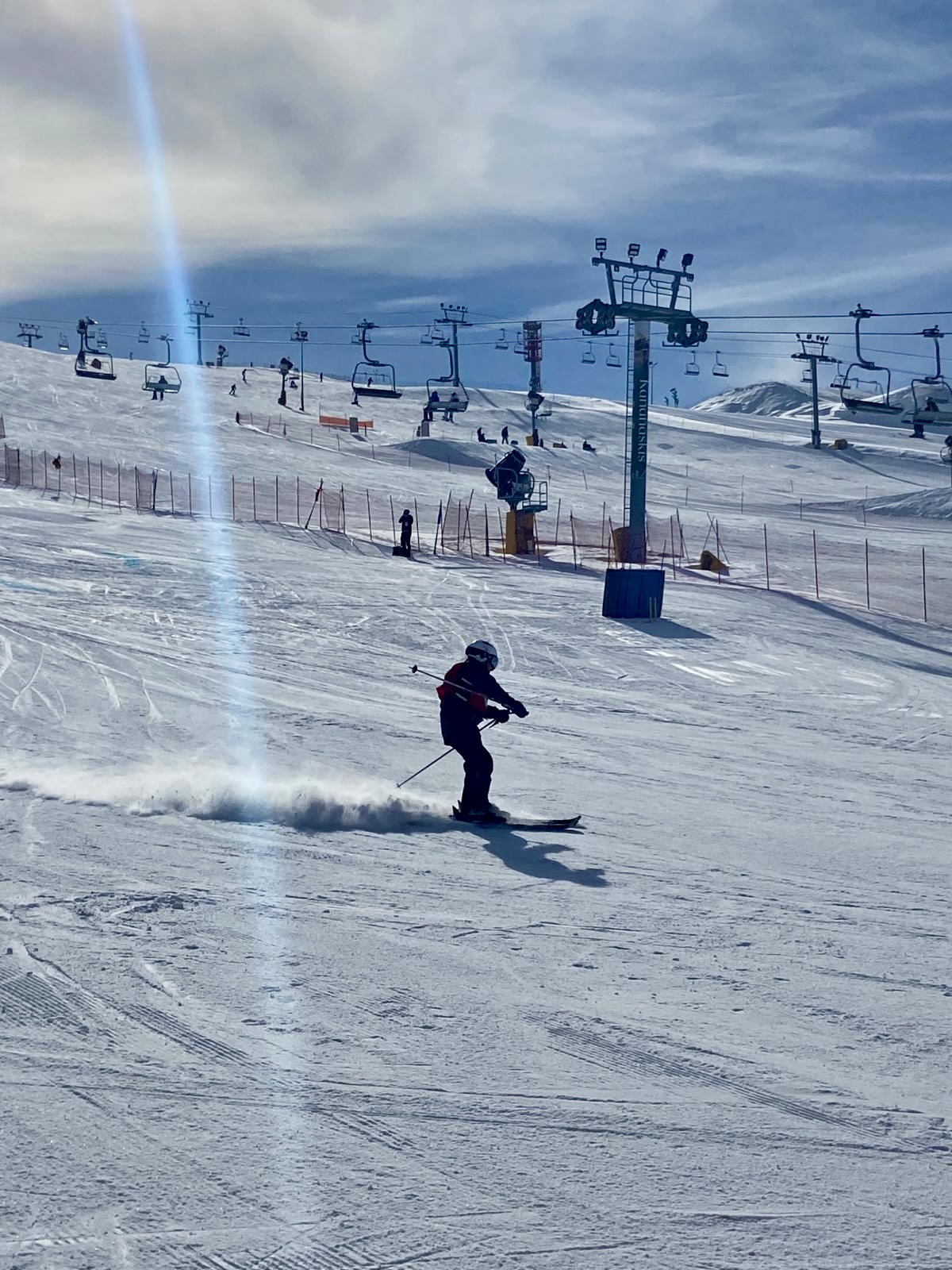 An action shot photo of an alpine skier going down the hill. The skier is a medium distance away from the camera and you can see the snow blowing behind their skis. In the distance behind the skier, you can see the ski lift and other skiers going down a different hill.