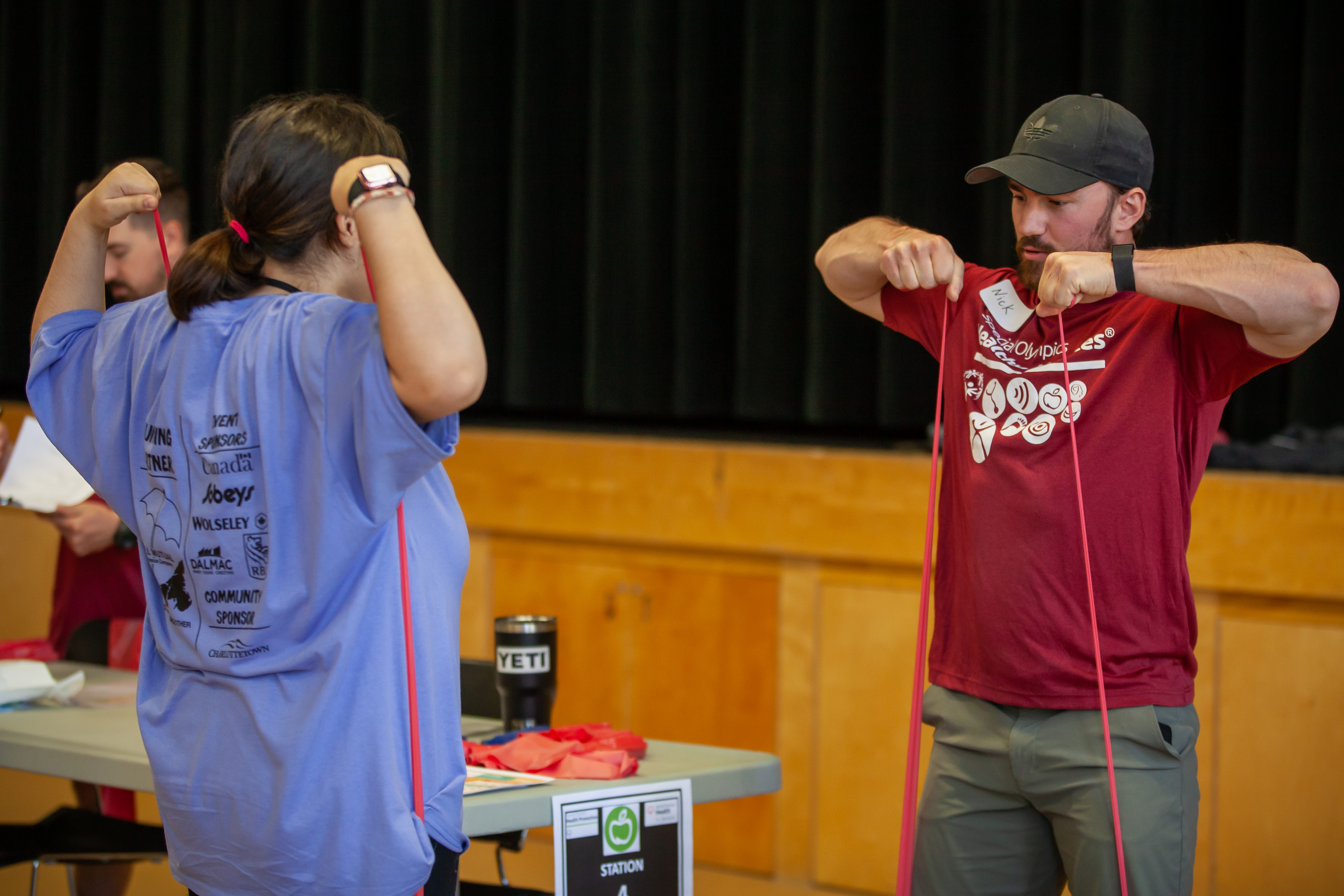 A volunteer instructor, is demonstrating and showing an athlete a stretching exercise using a red band. The athlete is following along with the instructor, with her back to the camera. A volunteer instructor, is demonstrating and showing an athlete a stretching exercise using a red band. The athlete is following along with the instructor, with her back to the camera.