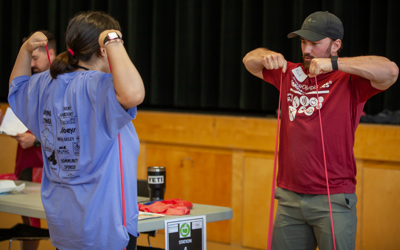 A volunteer instructor, is demonstrating and showing an athlete a stretching exercise using a red band. The athlete is following along with the instructor, with her back to the camera.