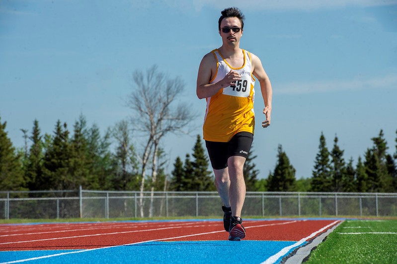 An athletics athlete is running down a track. He is the only athlete on the track. He is wearing a yellow tank top, with the number 459 pinned to it, as well as sunglasses, and black athletic shorts.