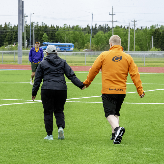 Two athletes holding hands walking on the field. The photo was captured from behind the athletes so you cannot see their faces. They are both wearing light rain jackets to prepare for the weather.