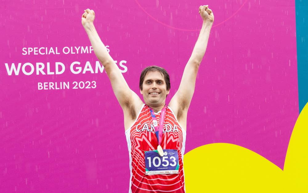Un athlète se tient sur la première marche du podium, les bras levés au-dessus de sa tête en signe de célébration, avec sa médaille d’or suspendue autour du cou.