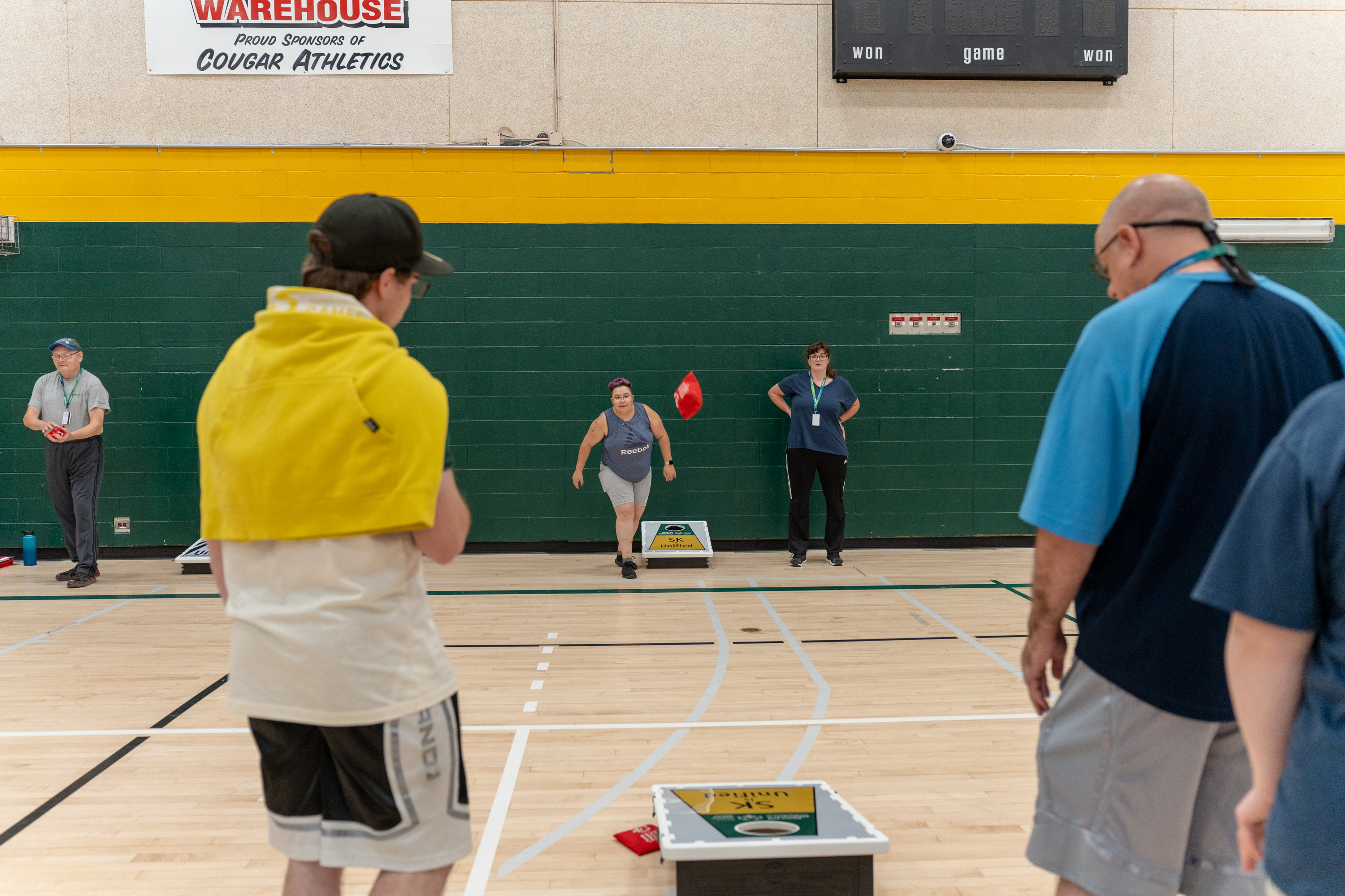 An action shot photo of people playing bean bag toss. In the distance you can see someone throwing a bean bag. The bean bag can be seen in midair coming closer to the camera. On the floor are the bean bag boards where the athletes are trying to toss the bean bags into. The game is being played inside a gymnasium. An action shot photo of people playing bean bag toss. In the distance you can see someone throwing a bean bag. The bean bag can be seen in midair coming closer to the camera. On the floor are the bean bag boards where the athletes are trying to toss the bean bags into. The game is being played inside a gymnasium.