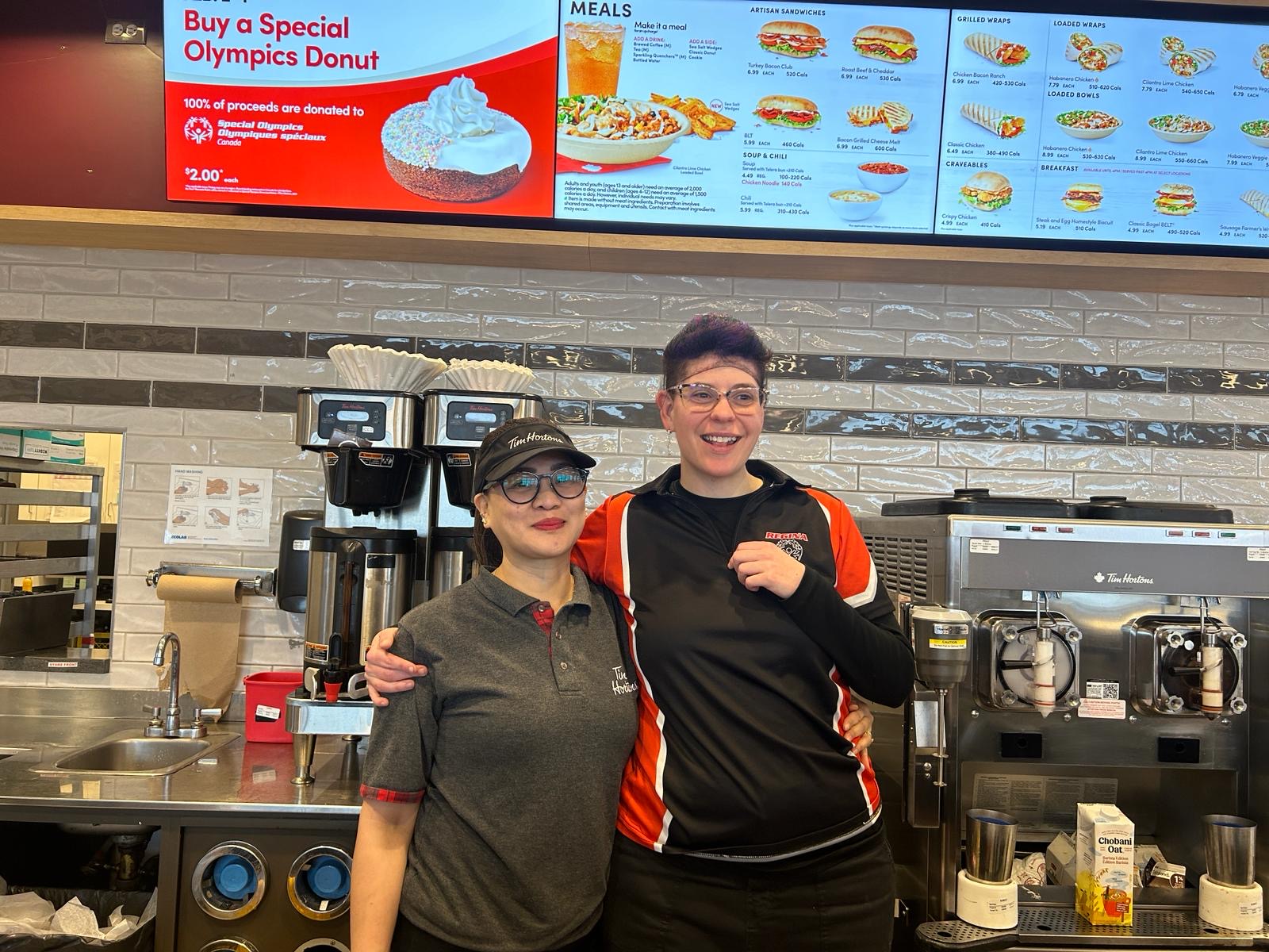 A female Special Olympics Saskatchewan athlete poses for the camera with a Tim Hortons employee. They are behind the Tim Hortons counter with their arms wrapped behind each other and they are smiling. Behind them are the display screens as well as coffee makers and equipment. A female Special Olympics Saskatchewan athlete poses for the camera with a Tim Hortons employee. They are behind the Tim Hortons counter with their arms wrapped behind each other and they are smiling. Behind them are the display screens as well as coffee makers and equipment.
