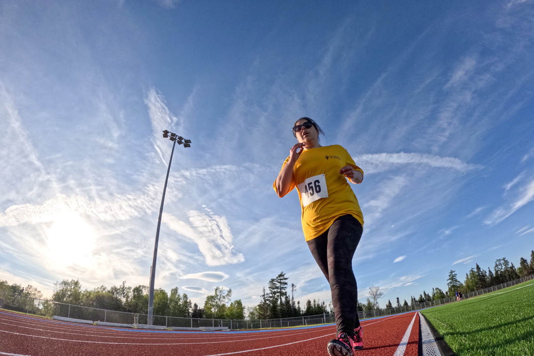 A slightly warped, low angled action shot photo of an athletics athlete running on the track. It is a bright, partly cloudy day and the athlete is wearing a yellow t-shirt with the number "456" pinned to the front of it. She is also wearing sunglasses and black and pink running cleats.