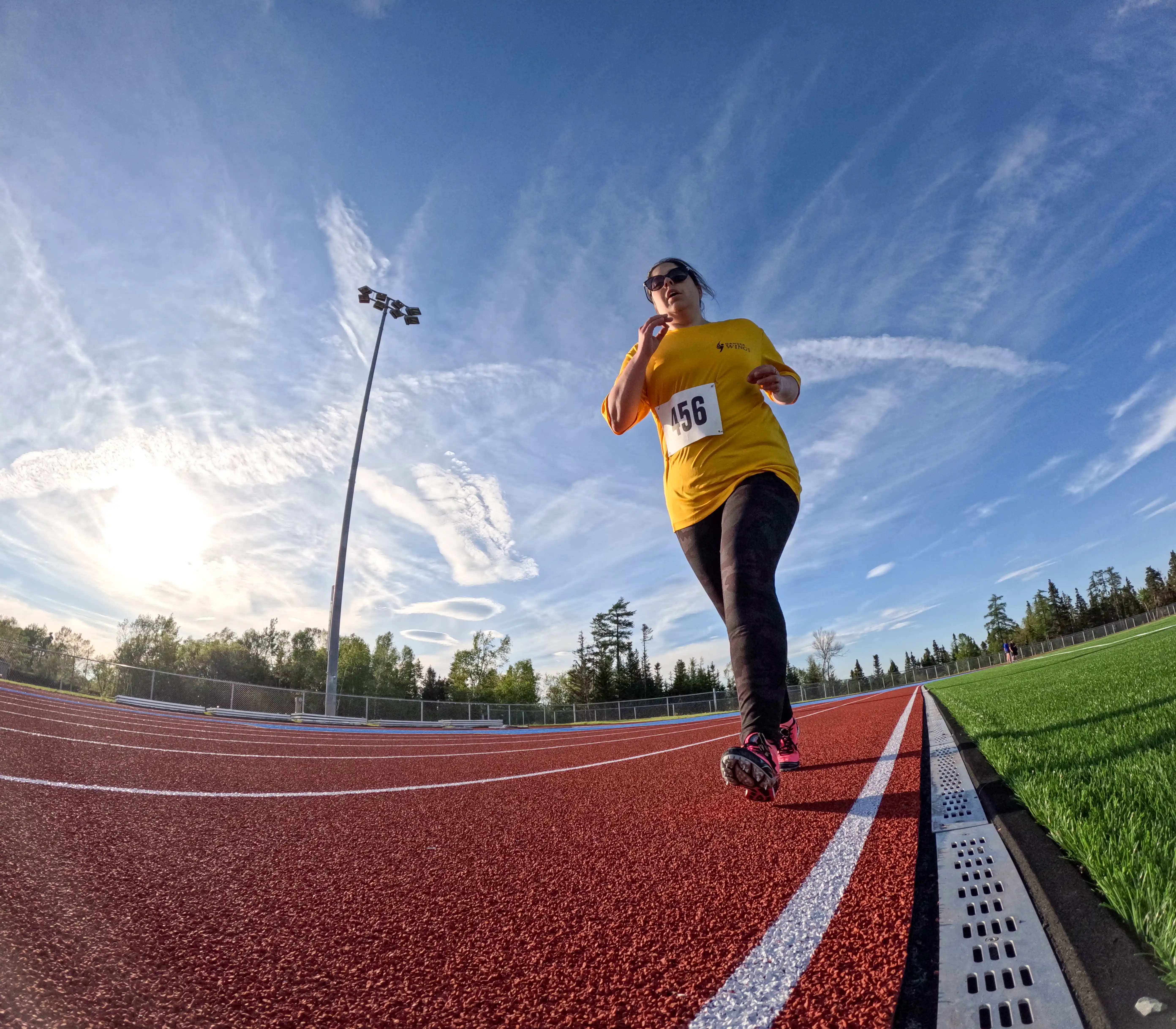 A slightly warped, low angled action shot photo of an athletics athlete running on the track. It is a bright, partly cloudy day and the athlete is wearing a yellow t-shirt with the number 