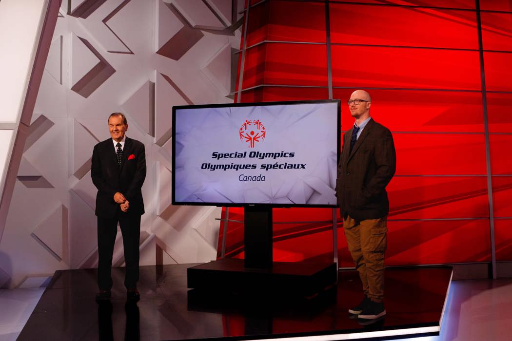 Deux hommes debout, tous deux en costume. Entre eux se trouve un écran de télévision avec le logo des Olympiques spéciaux du Canada. 