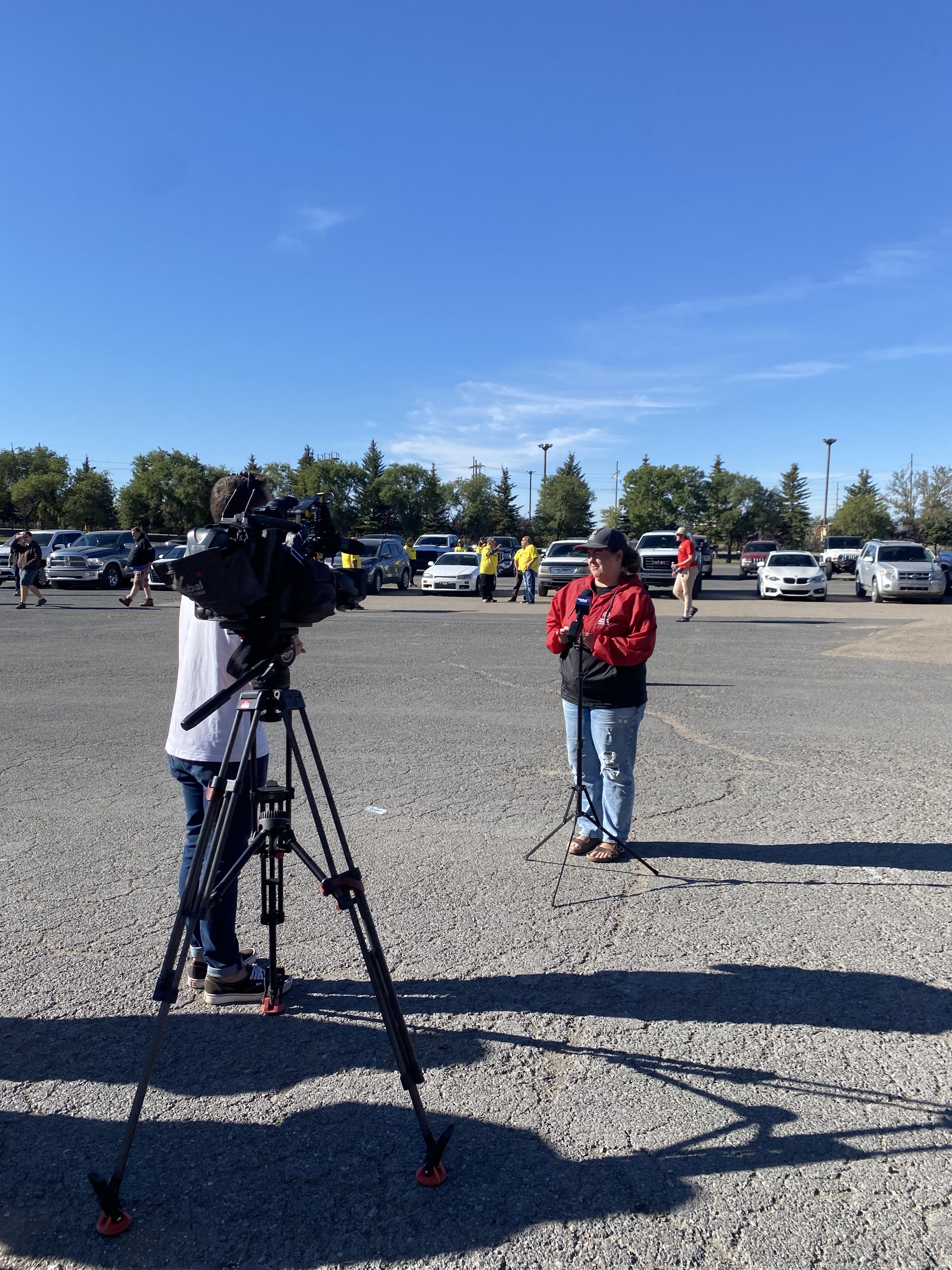 A medium shot photo of a camera on a tripod. There is an individual behind the camera, but the camera is obstructing him from view. There is also an individual in front of the camera. You can see their full body and they are wearing a red jacket with jeans. A medium shot photo of a camera on a tripod. There is an individual behind the camera, but the camera is obstructing him from view. There is also an individual in front of the camera. You can see their full body and they are wearing a red jacket with jeans.