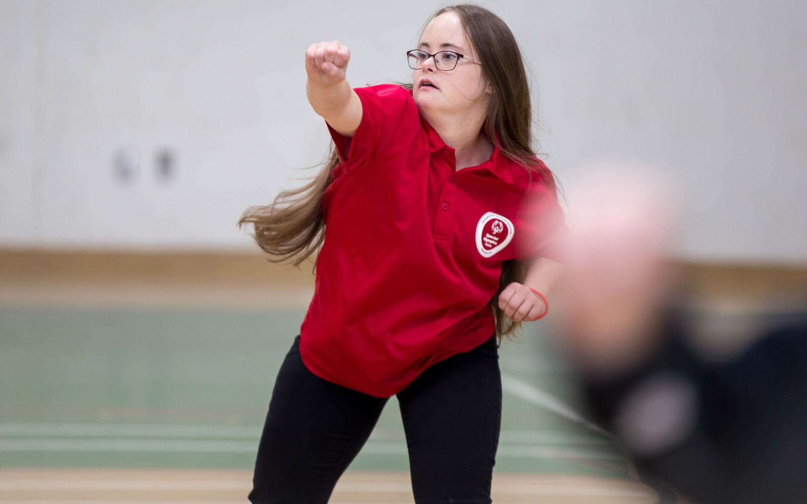 A photo of an athlete in motion, focusing on her FUNfitness activity. She is the only person in focus, the rest of the photo is blurry. She is wearing a red Special Olympics collared shirt, and glasses.