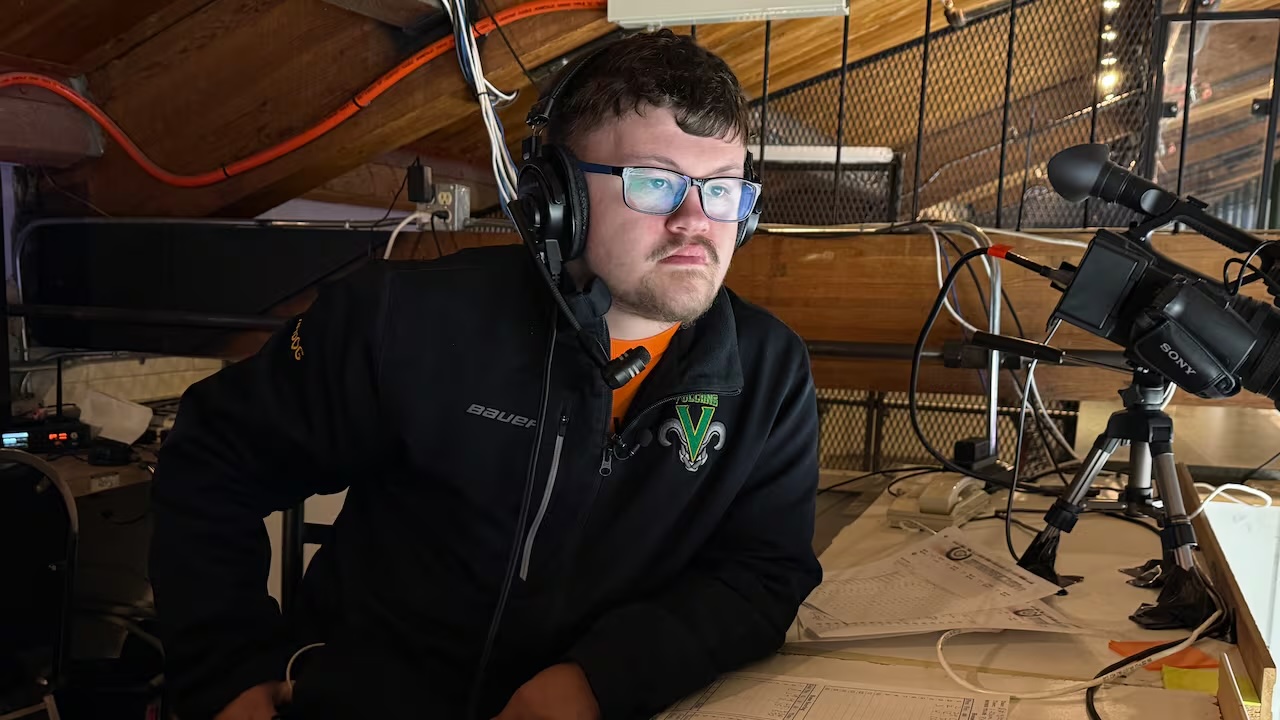 Tanner has headphones on and is sitting behind a microphone in a hockey arena. 