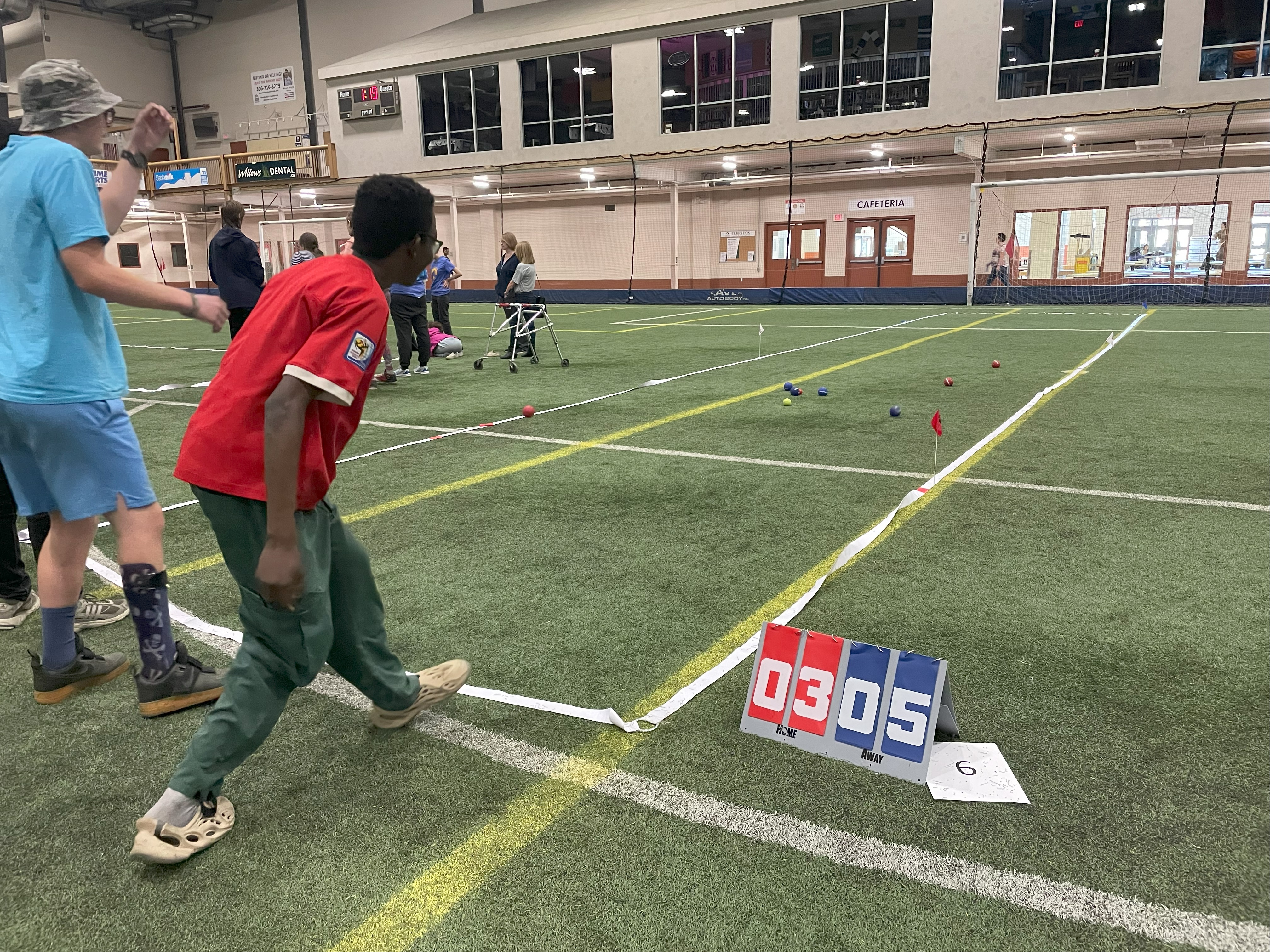An action shot photo of an athlete playing bocce. He is watching as the ball he threw lands on the grass in front of him. You can only see the athlete from behind. On the field, you can see in the distance the other bocce balls that are in place. Next to the space defined for the game, is a scoreboard reading An action shot photo of an athlete playing bocce. He is watching as the ball he threw lands on the grass in front of him. You can only see the athlete from behind. On the field, you can see in the distance the other bocce balls that are in place. Next to the space defined for the game, is a scoreboard reading