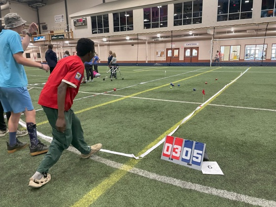 An action shot photo of an athlete playing bocce. He is watching as the ball he threw lands on the grass in front of him. You can only see the athlete from behind. On the field, you can see in the distance the other bocce balls that are in place. Next to the space defined for the game, is a scoreboard reading "03" for the red team and "05" for the blue team.