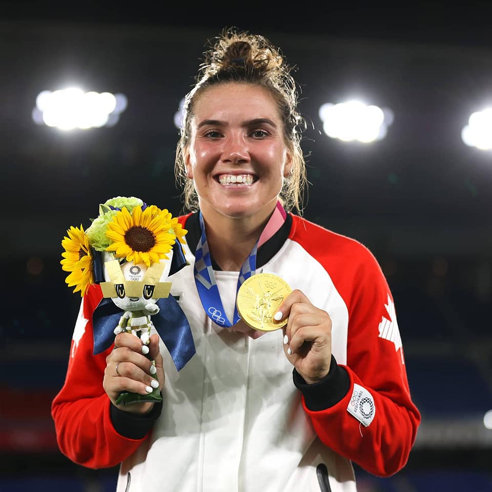Headshot of a woman with hair in a topknot. She is wearing a Team Canada zip up and holding a bouquet of sunflowers. Around her neck is a gold medal. Headshot of a woman with hair in a topknot. She is wearing a Team Canada zip up and holding a bouquet of sunflowers. Around her neck is a gold medal.