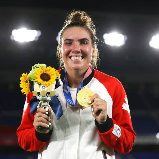 Gros plan sur le visage d'une femme aux cheveux relevés en chignon. Elle porte une veste zippée de l'équipe canadienne et tient un bouquet de tournesols. Elle porte une médaille d'or autour du cou.
