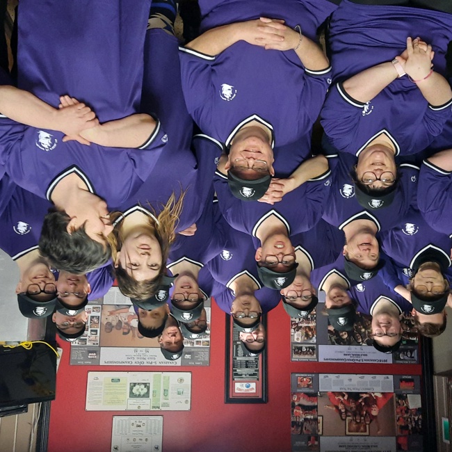 A group photo where everyone is wearing matching purple jerseys. There are 5 rows of people of mixed genders and ages, and everyone is smiling for the camera.
