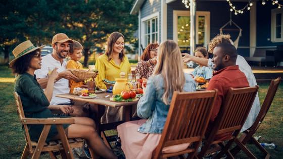 Group of people sitting outside at a picnic table, with table covered in food and drinks. 