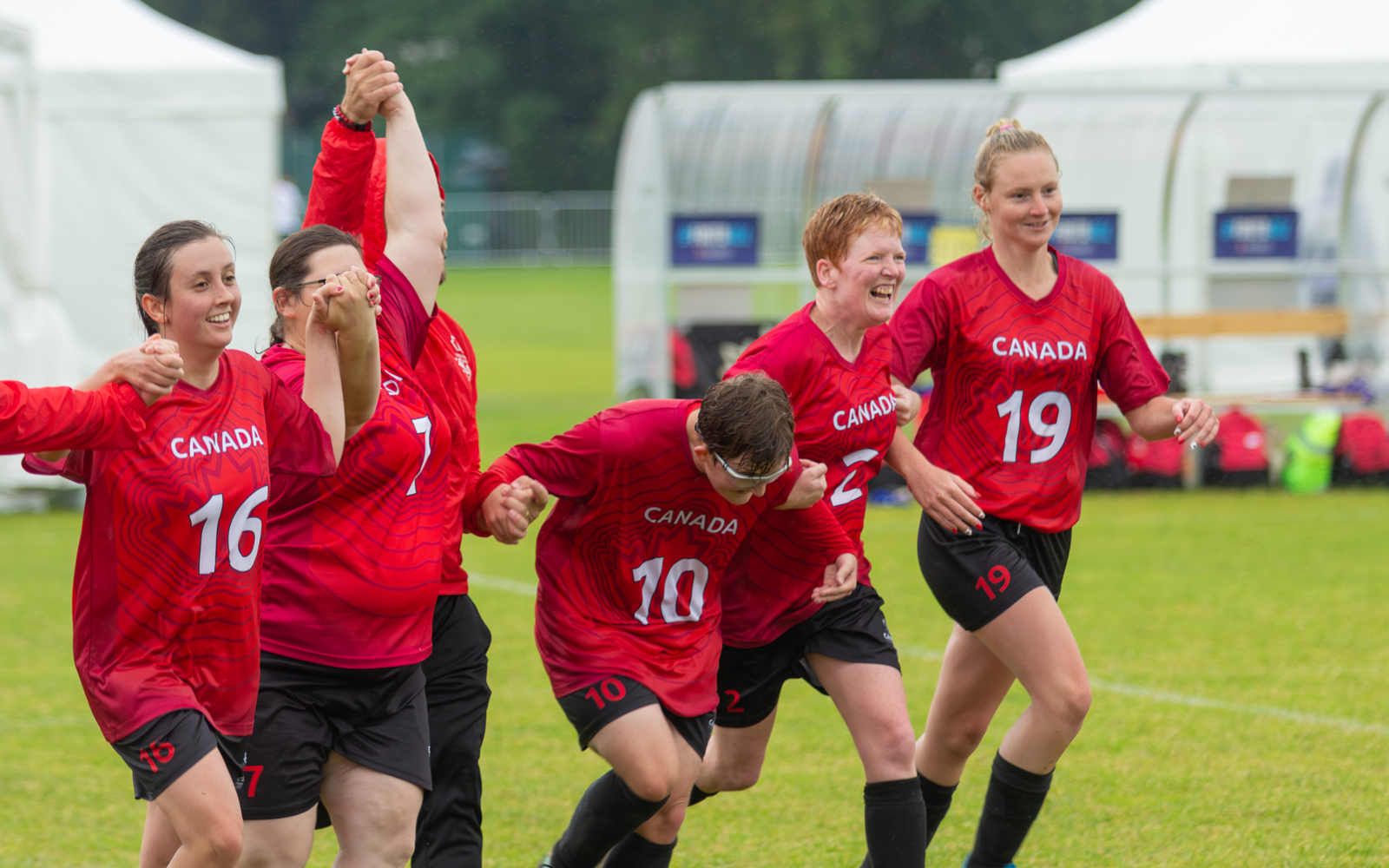 6 athlètes féminines vêtues de maillots de football canadiens rouges et de shorts noirs se tenant par la main, souriant et se dirigeant vers la caméra. Toutes semblent avoir participé à un match de football.