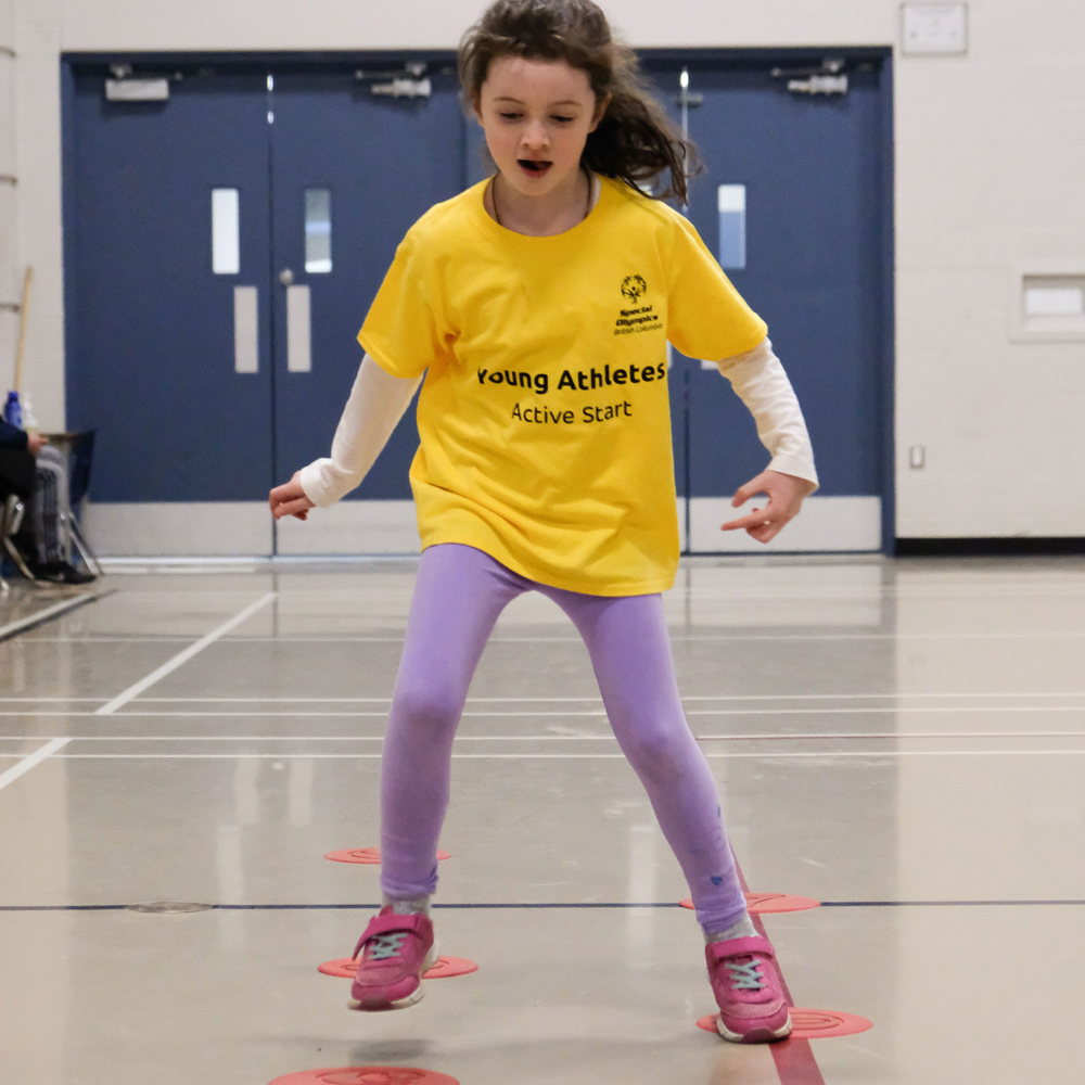 A young girl with intellectual disabilities jumps between markers on the floor looking very focused.