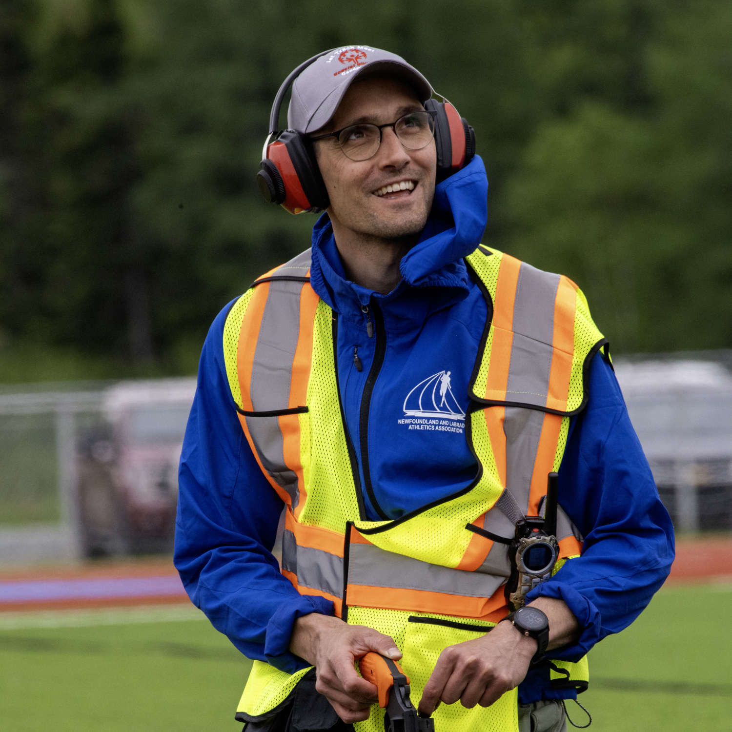 An image of a race starter, on his knees, reloading the starting gun. He is wearing a high visibility vest as well as large red headphones to protect his ears. He is looking up and to the right of the camera, smiling and listening to someone who does not appear in the photo.