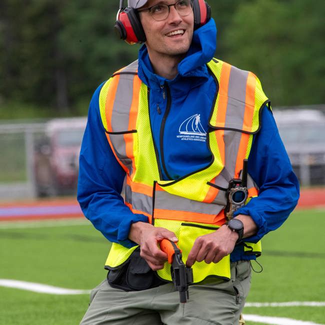 An image of a race starter, on his knees, reloading the starting gun. He is wearing a high visibility vest as well as large red headphones to protect his ears. He is looking up and to the right of the camera, smiling and listening to someone who does not appear in the photo.