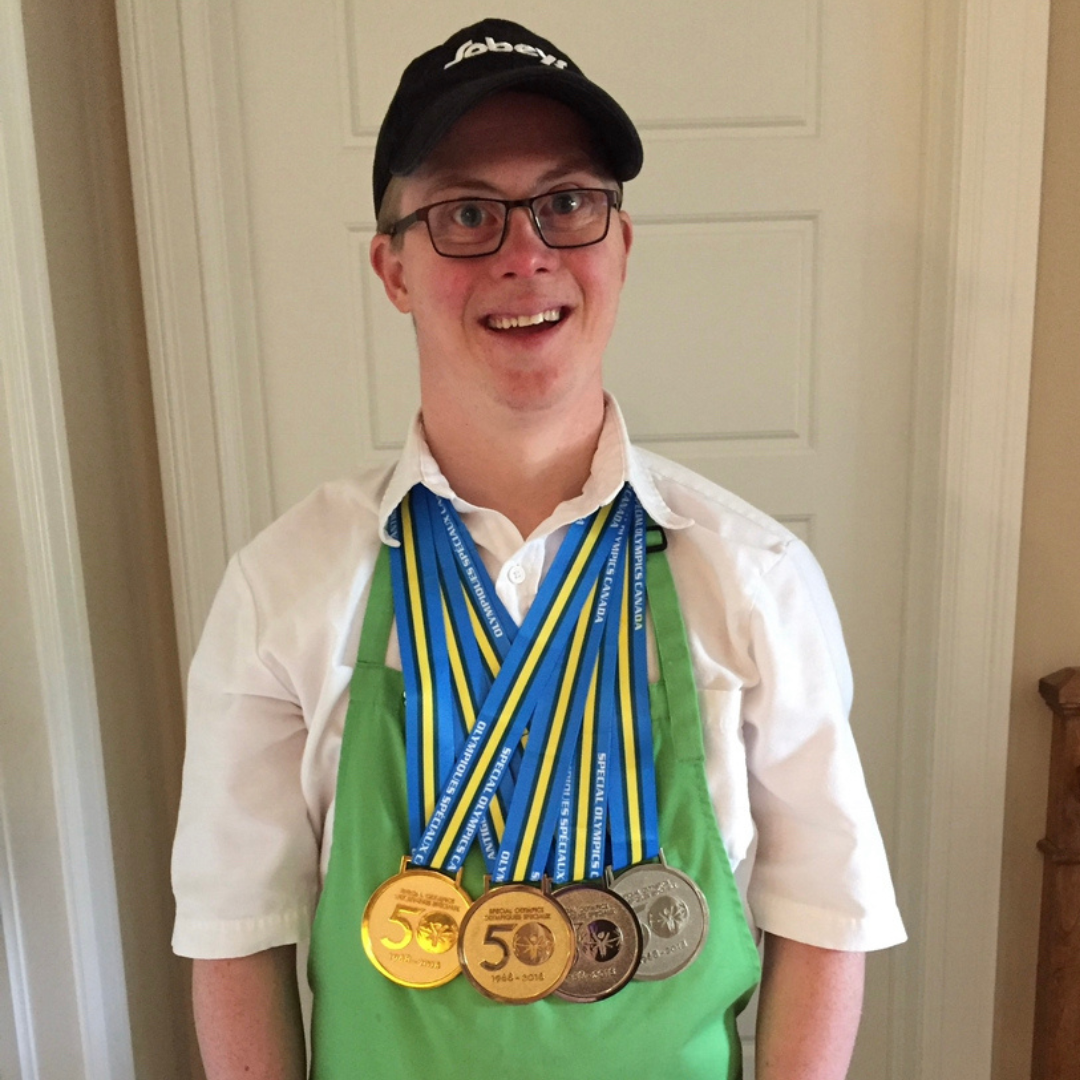 Young man with a white shirt, green apron and black hat smiles at the camera. He has dark glasses and is wearing 4 meals around his neck. 