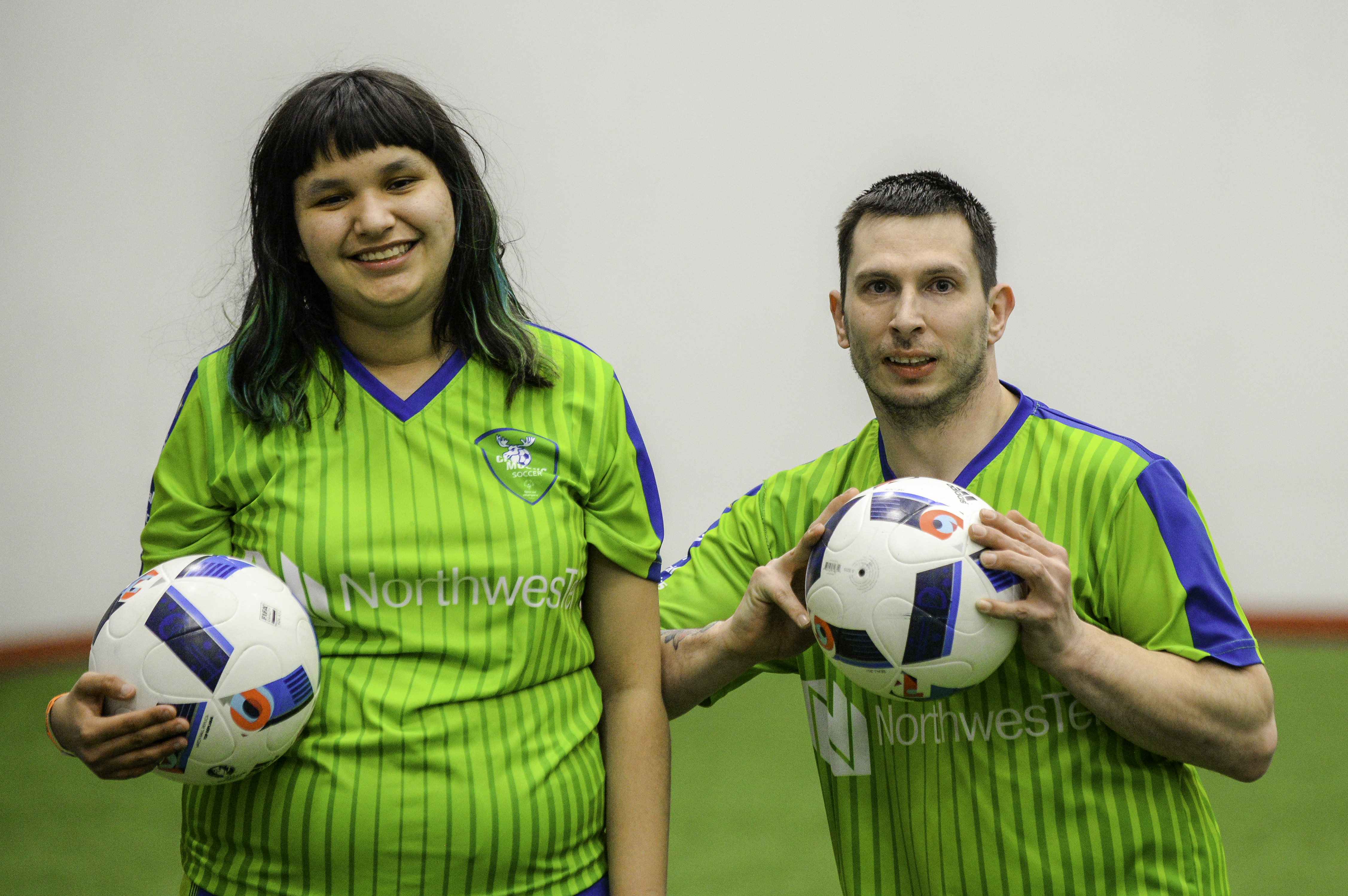 A close up of two soccer players smiling at the camera and posing with a soccer ball. Both athletes are wearing bright green soccer jerseys and holding white and blue soccer balls.
