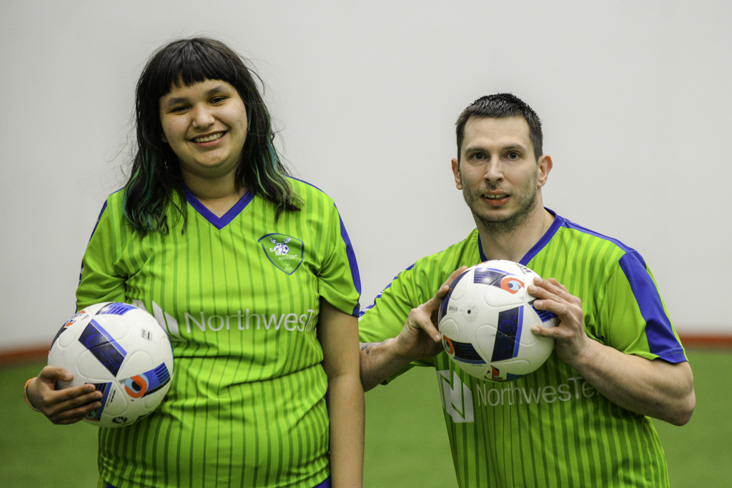 A close up of two soccer players smiling at the camera and posing with a soccer ball. Both athletes are wearing bright green soccer jerseys and holding white and blue soccer balls.