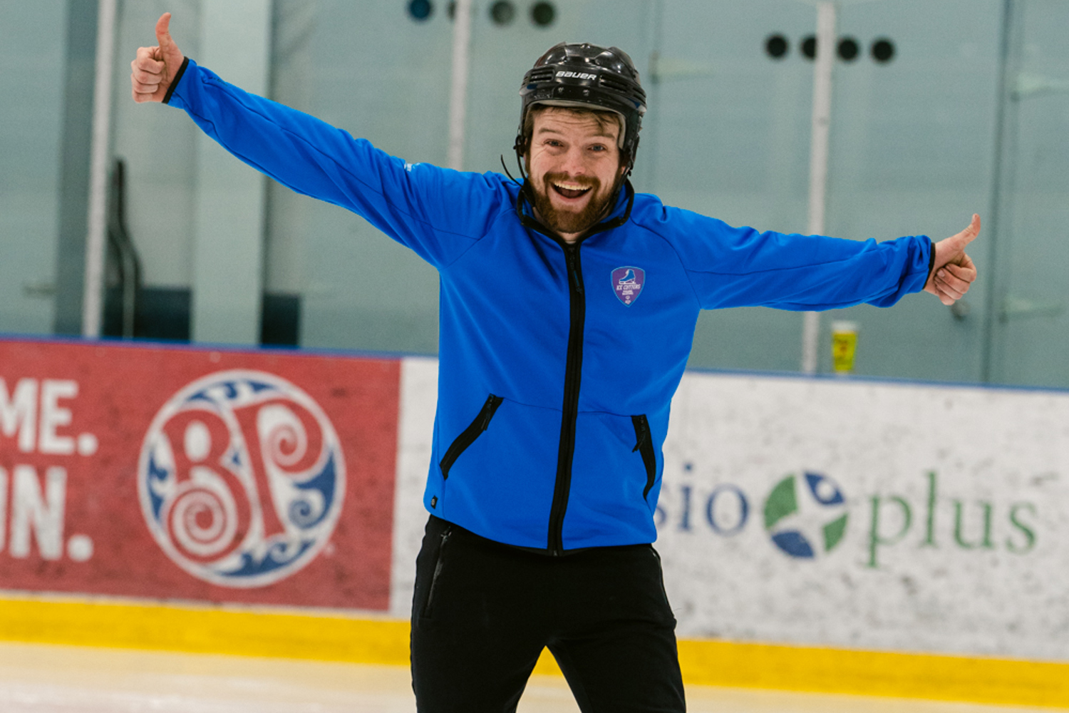 A male figure skater happily poses for the camera. His arms are stretched out wide and he is giving two thumbs up. He also has one leg out slightly with the edge of his skate on the ice. He is wearing a blue sweater and a black helmet.