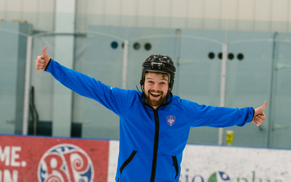 A male figure skater happily poses for the camera. His arms are stretched out wide and he is giving two thumbs up. He also has one leg out slightly with the edge of his skate on the ice. He is wearing a blue sweater and a black helmet.
