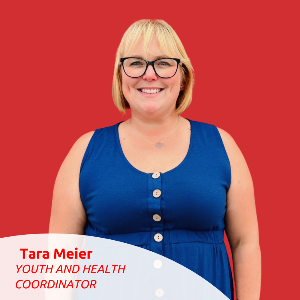 Portrait Photo of Tara Meier. She is standing against a red wall and smiling for the camera.