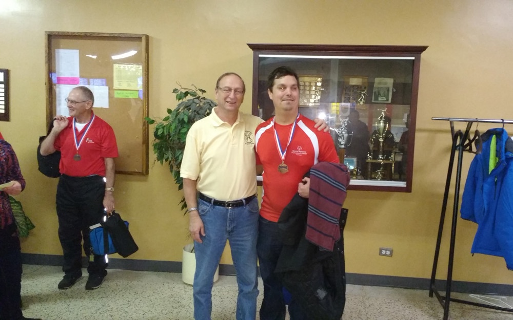 Two individuals are posing and smiling for the camera. The athlete on the right is wearing a red jersey and has a medal around his neck. The individual on the left is wearing a yellow collared t-shirt and has his arm around the back of the athlete. They are standing in a room, and are in front of a trophy case.