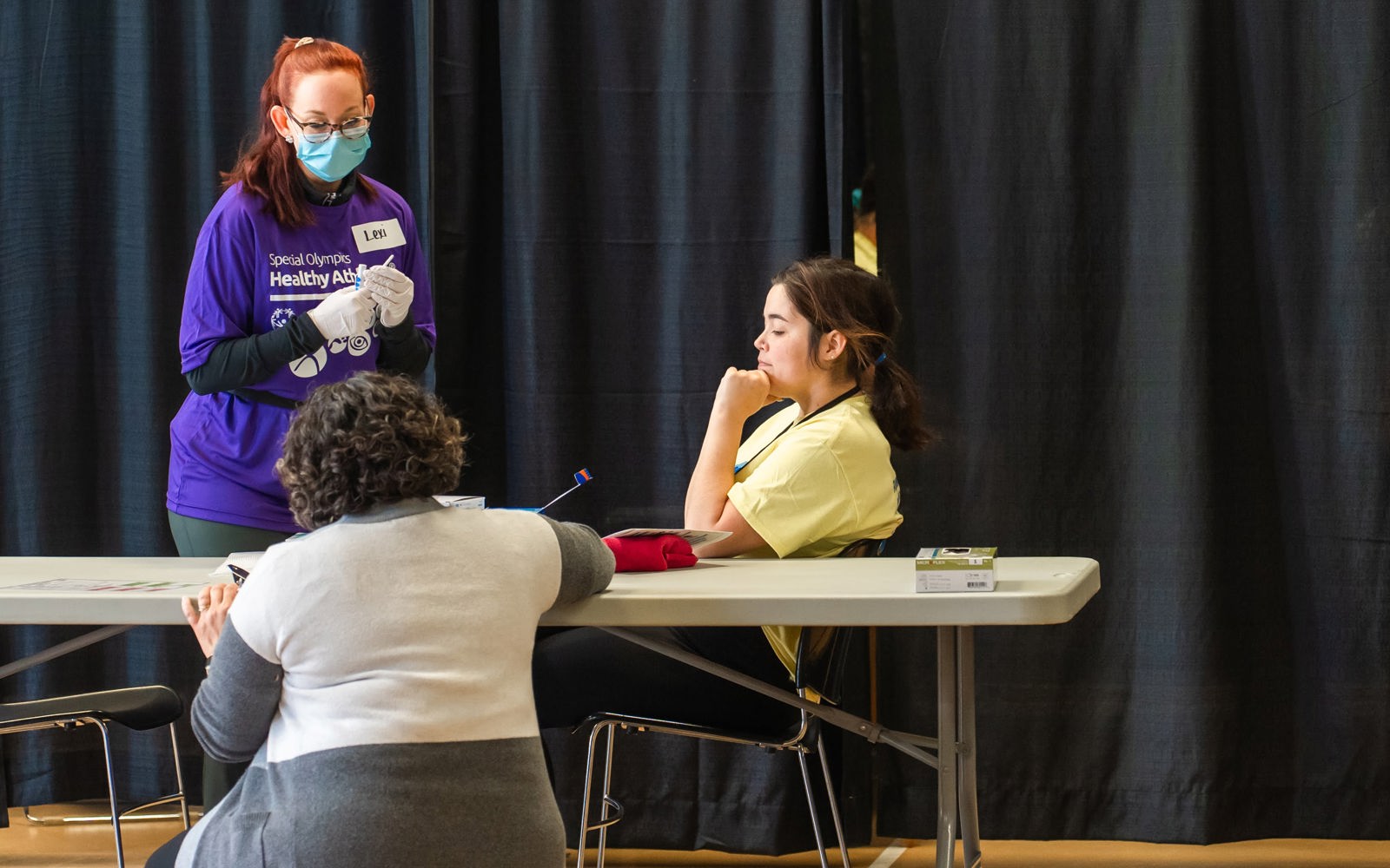 women in a purple shirt wearing a mask talks to two other women, one in a yellow shirt and one in a grey sweater.