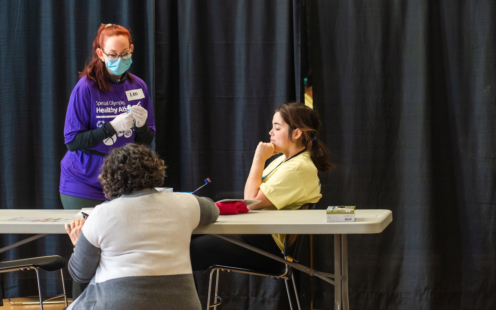 women in a purple shirt wearing a mask talks to two other women, one in a yellow shirt and one in a grey sweater. 
