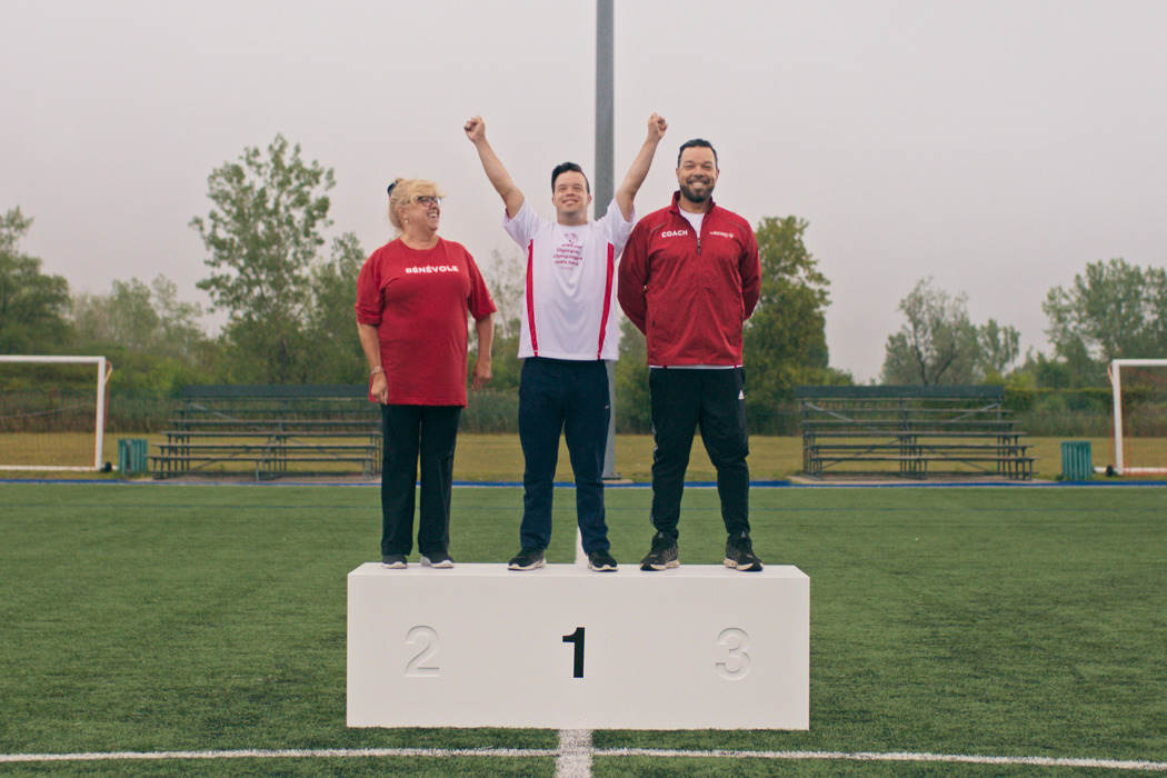 Trois personnes se tiennent sur un podium dans un terrain de football. La femme à gauche est en haut du placement 2 et porte un maillot rouge. L'homme au milieu est en haut du placement 1 et a les bras en l'air. L'homme à droite est en haut du placement 3 et porte un pull rouge. 