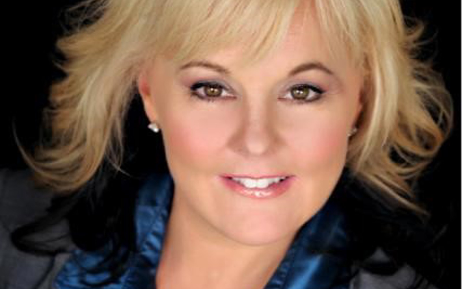 Portrait d'une femme aux cheveux blonds vêtue d'une veste grise et d'une chemise bleue.