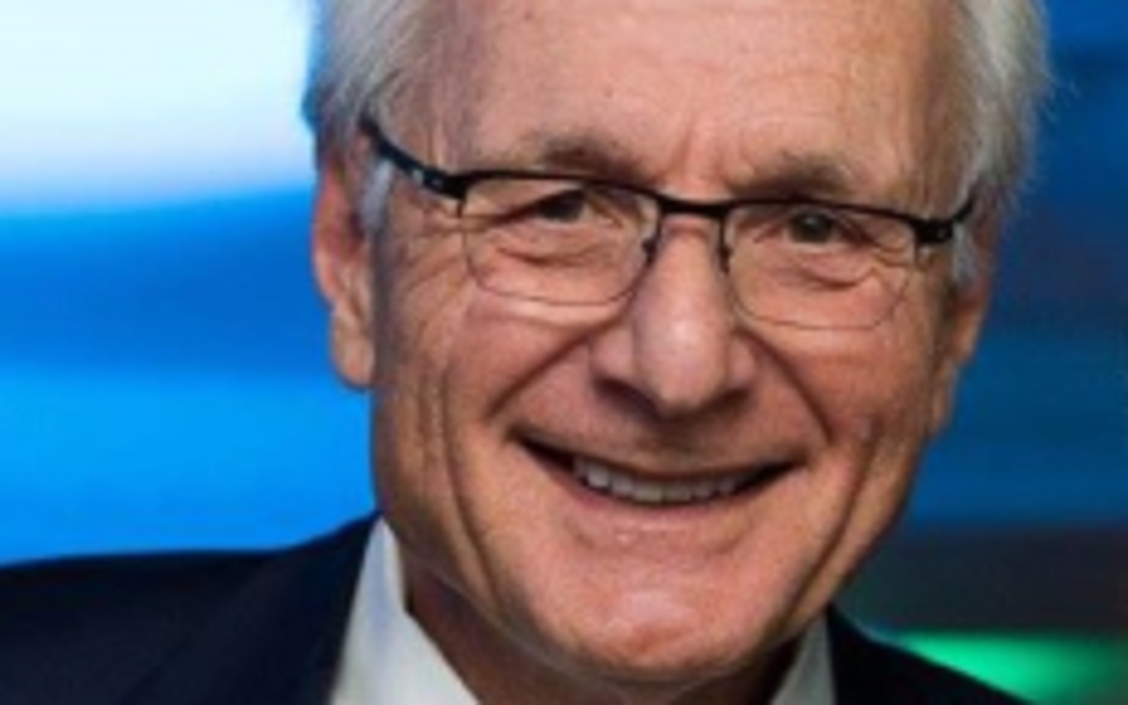 Man in black suit, white shirt and blue tie with glasses smiling at camera.