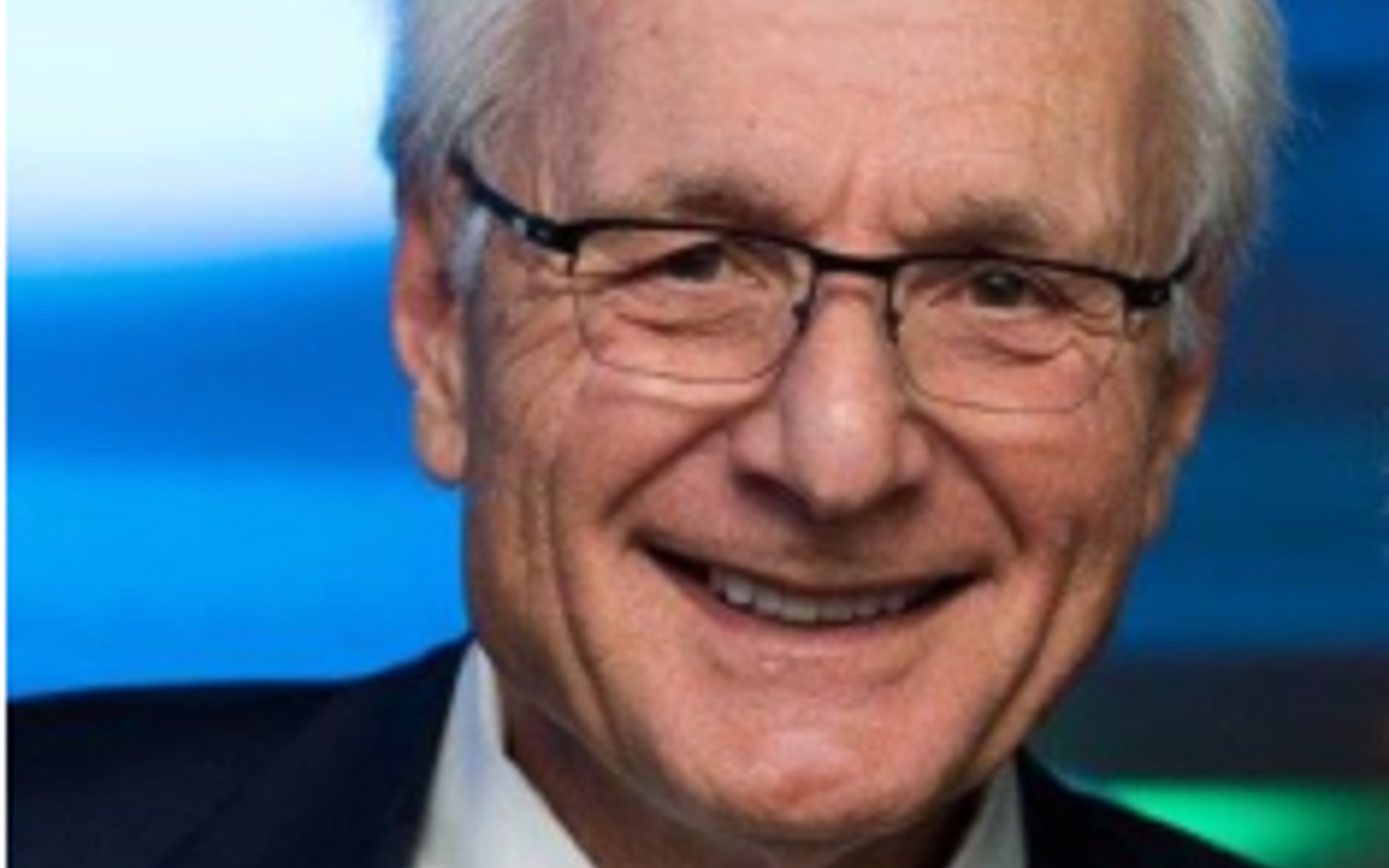Man in black suit, white shirt and blue tie with glasses smiling at camera. 