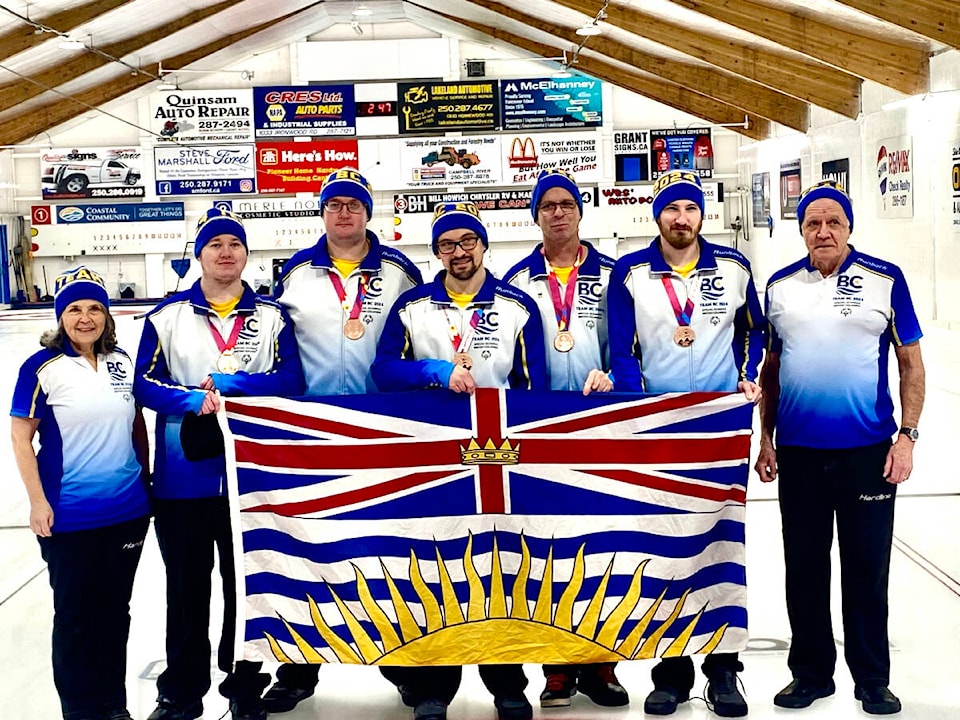 7 people standing in a line. All dressed in Special Olympics British Columbia curling uniforms. Holding the BC flag. 5 of them have medals around thier necks. 