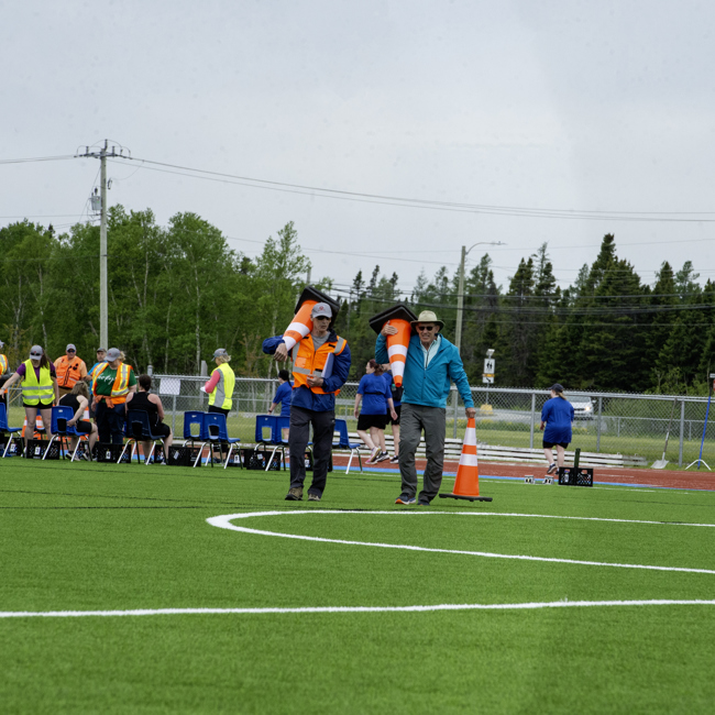Two individuals are walking on a field holding large, orange, pylons. The individual on the left is wearing a high visibility vest and is holding one pylon while the individual on the right is wearing a blue jacket and holding two pylons. In the background you can see other officials and volunteers in a group.