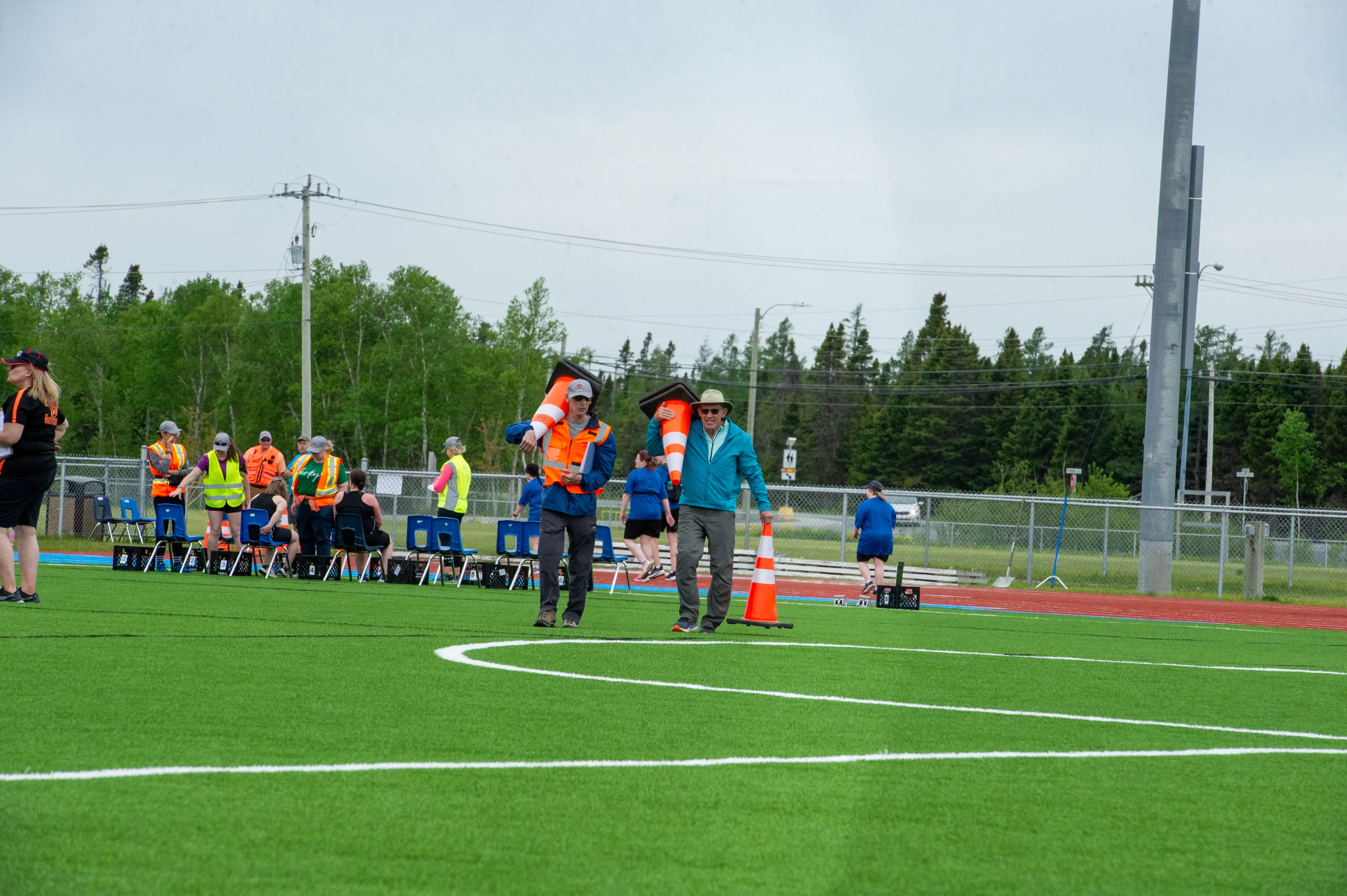 Two individuals are walking on a field holding large, orange, pylons. The individual on the left is wearing a high visibility vest and is holding one pylon while the individual on the right is wearing a blue jacket and holding two pylons. In the background you can see other officials and volunteers in a group. Two individuals are walking on a field holding large, orange, pylons. The individual on the left is wearing a high visibility vest and is holding one pylon while the individual on the right is wearing a blue jacket and holding two pylons. In the background you can see other officials and volunteers in a group.