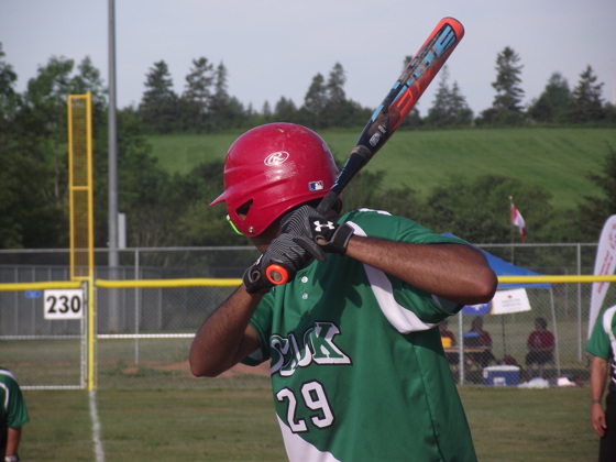 A close up image of an athlete playing baseball. They have their face turned away from the camera as they are focusing on their game. They are swinging the baseball bat back in preparation to hit the ball. They are wearing a green baseball jersey.